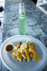 A close-up view of a plate with three tacos topped with cheese, vegetables, and meat alongside a small cup of green salsa. The background features a glass bottle of Mundet apple soda on a marble-like counter.