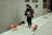 A person dressed warmly is walking three small dogs on a sidewalk. The dogs are all wearing colorful jackets; one is in a red jacket, another in a red one with yellow spots, and the third in a dark jacket. The person is also holding a smartphone, possibly checking messages or navigation, and has earphones connected. The background is an urban setting with a gray brick wall.
