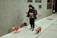 A person dressed warmly is walking three small dogs on a sidewalk. The dogs are all wearing colorful jackets; one is in a red jacket, another in a red one with yellow spots, and the third in a dark jacket. The person is also holding a smartphone, possibly checking messages or navigation, and has earphones connected. The background is an urban setting with a gray brick wall.