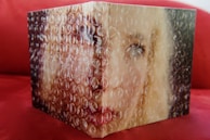 A partially visible portrait of a person obscured by bubble wrap, with a focus on the eyes and blonde hair. The background is a red surface, likely a couch or similar furniture.