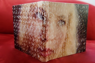 A partially visible portrait of a person obscured by bubble wrap, with a focus on the eyes and blonde hair. The background is a red surface, likely a couch or similar furniture.