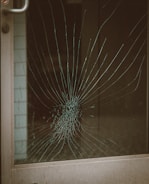 A glass door with a large spider web crack at the center, indicating significant impact. The crack radiates outward in a pattern typical of shattered glass. The metal door frame is visible on the left side.