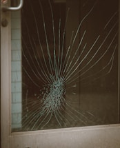 A glass door with a large spider web crack at the center, indicating significant impact. The crack radiates outward in a pattern typical of shattered glass. The metal door frame is visible on the left side.