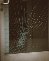 A glass door with a large spider web crack at the center, indicating significant impact. The crack radiates outward in a pattern typical of shattered glass. The metal door frame is visible on the left side.