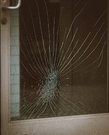 A glass door with a large spider web crack at the center, indicating significant impact. The crack radiates outward in a pattern typical of shattered glass. The metal door frame is visible on the left side.