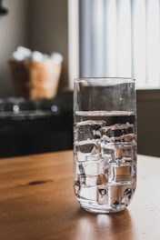 A clear glass filled with ice cubes sits on a wooden table. In the background, a blurred basket is holding white objects, and a window with vertical blinds allows soft light into the room.