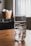 A clear glass filled with ice cubes sits on a wooden table. In the background, a blurred basket is holding white objects, and a window with vertical blinds allows soft light into the room.