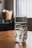 A clear glass filled with ice cubes sits on a wooden table. In the background, a blurred basket is holding white objects, and a window with vertical blinds allows soft light into the room.
