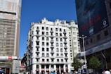 High-rise building with white façade and numerous balconies in an urban setting. On the left, another building is covered in scaffolding and netting, indicating construction work. A large advertisement poster is displayed on the building to the right. Pedestrians can be seen at the bottom, walking and standing near street signs and a Metro entrance. The sky is clear and blue.