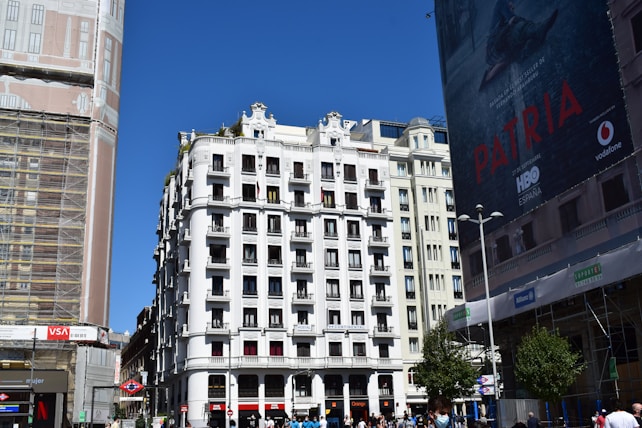 High-rise building with white façade and numerous balconies in an urban setting. On the left, another building is covered in scaffolding and netting, indicating construction work. A large advertisement poster is displayed on the building to the right. Pedestrians can be seen at the bottom, walking and standing near street signs and a Metro entrance. The sky is clear and blue.