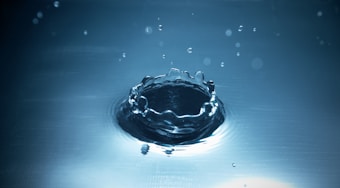 A close-up view of a water droplet impact, creating a crown-like splash with ripples spreading outward on a smooth surface. Smaller droplets are suspended above, adding to the sense of motion and fluidity.