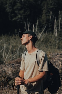 A young man stands outdoors with his eyes closed, facing the sunlight. He is wearing a beige t-shirt, a black cap, and is carrying a backpack. The background is a blurred forest scene, creating a serene atmosphere.