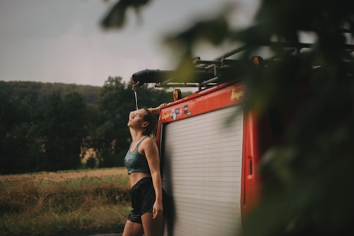 A person is leaning against a red fire truck in an outdoor, rural setting. They appear to be cooling off, with water possibly spraying or dripping onto their face from the truck. The person is dressed in athletic clothing, suggesting they might be cooling down after exercise. The background features lush greenery and a field, creating a serene and natural environment.
