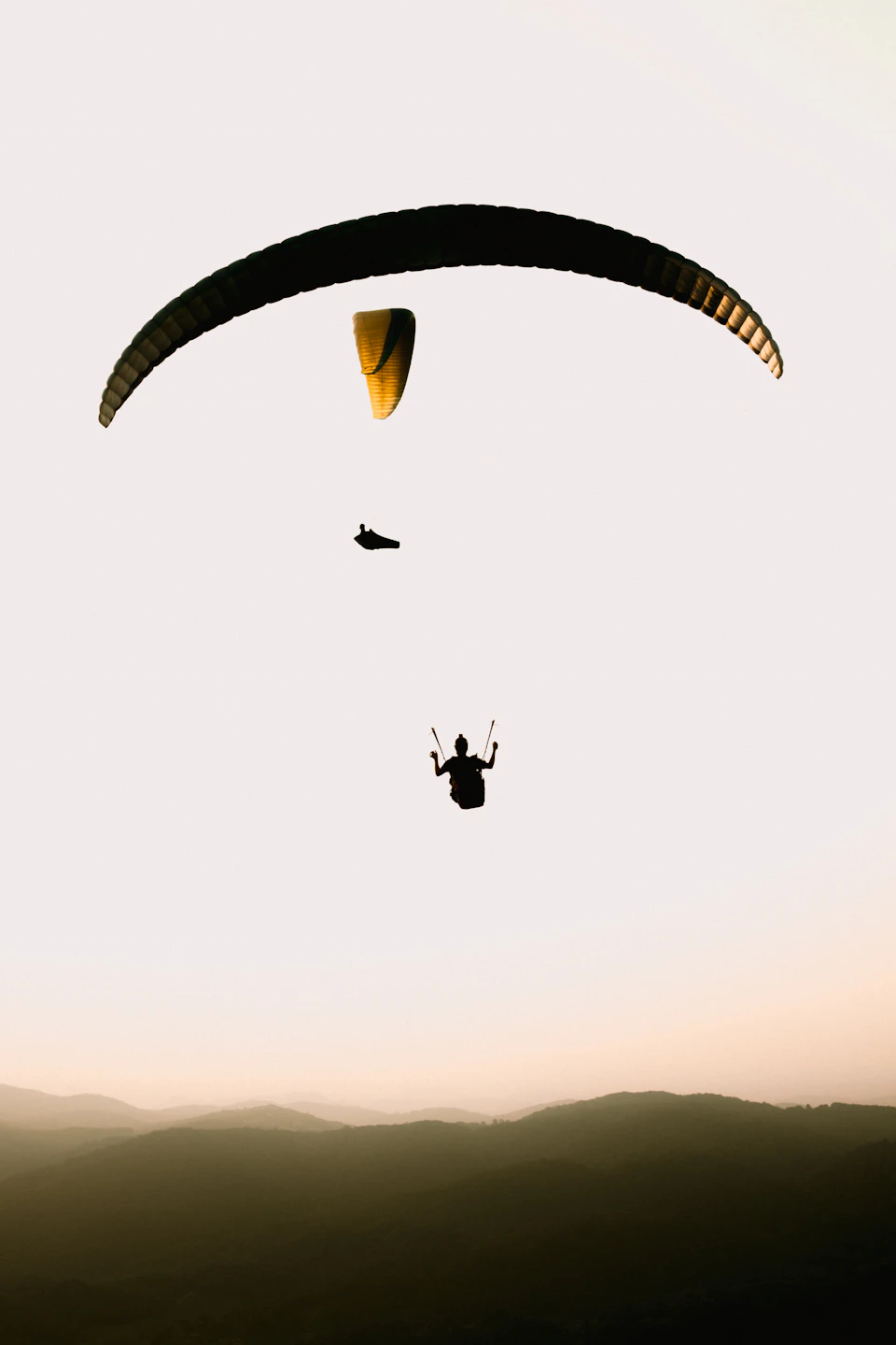 Two people paraglide over a mountain range during sunset with orange and yellow hues