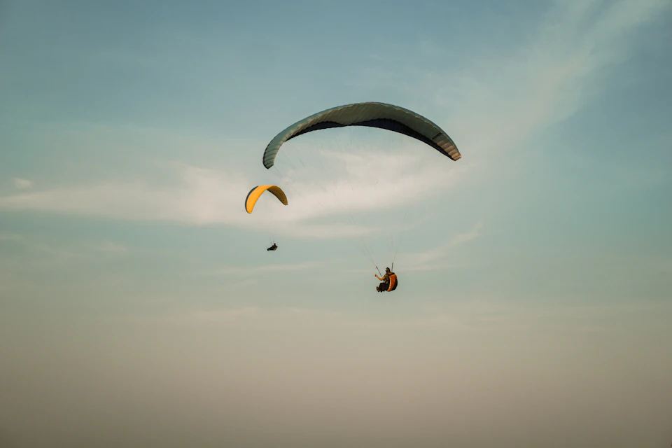 The image depicts three paragliders soaring through a clear blue sky with fluffy white clouds. The first paraglider is flying towards the left side of the frame, while the second and third are positioned more centrally, creating an interesting visual dynamic. The paragliders' colorful parachutes contrast beautifully against the azure backdrop, adding depth to the scene.