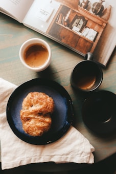A cozy scene with a flaky croissant on a blue plate, accompanied by two cups of coffee. An open book displays warm-toned photography or artwork, suggesting a relaxed atmosphere. The setup rests on a soft cloth atop a rustic wooden table, creating a warm and inviting setting.