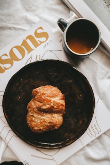 A croissant sits on a dark plate placed on top of a magazine with a partially visible title in bold, golden letters. Next to it, there is a dark ceramic cup filled with coffee. The background is a white cloth, contributing to the cozy and inviting atmosphere.