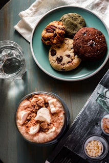 A blue plate holds an assortment of four cookies with different textures and ingredients, including chocolate chips and nuts. Next to the plate is a clear glass of water and a frothy beverage topped with marshmallows and crumb toppings. A magazine or book is partially visible on the table surface, which is wooden and has a dark stain.