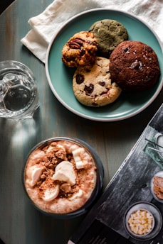 A blue plate holds an assortment of four cookies with different textures and ingredients, including chocolate chips and nuts. Next to the plate is a clear glass of water and a frothy beverage topped with marshmallows and crumb toppings. A magazine or book is partially visible on the table surface, which is wooden and has a dark stain.