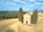 A small stone chapel is surrounded by tall evergreen trees in a vast open field. The landscape is characterized by rolling hills, with patches of green forests and yellowed grasslands. A few people are walking along a pathway near the chapel, and the sky is partly cloudy with a bright blue color.