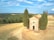 A small stone chapel is surrounded by tall evergreen trees in a vast open field. The landscape is characterized by rolling hills, with patches of green forests and yellowed grasslands. A few people are walking along a pathway near the chapel, and the sky is partly cloudy with a bright blue color.