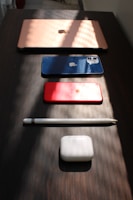 A neatly arranged setup of electronic devices on a dark wooden surface includes a rose gold laptop, a blue smartphone, a red tablet, a white stylus, and white earbud charging case. Soft, angled lighting creates patterns of shadows and highlights across the scene, accentuating the sleek design of the gadgets.