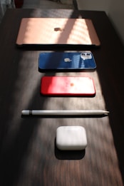 A neatly arranged setup of electronic devices on a dark wooden surface includes a rose gold laptop, a blue smartphone, a red tablet, a white stylus, and white earbud charging case. Soft, angled lighting creates patterns of shadows and highlights across the scene, accentuating the sleek design of the gadgets.