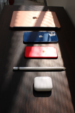 A neatly arranged setup of electronic devices on a dark wooden surface includes a rose gold laptop, a blue smartphone, a red tablet, a white stylus, and white earbud charging case. Soft, angled lighting creates patterns of shadows and highlights across the scene, accentuating the sleek design of the gadgets.