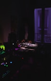 A dimly lit room with a person operating DJ equipment. Purple and blue lighting creates a moody atmosphere, and various electronic devices are visible, adding to the ambiance of a night-time music setup.