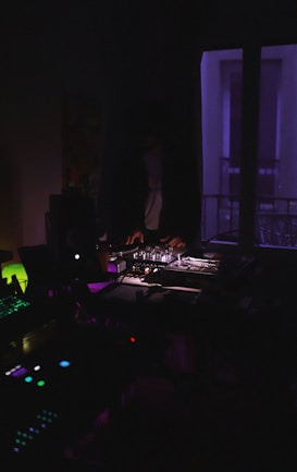 A dimly lit room with a person operating DJ equipment. Purple and blue lighting creates a moody atmosphere, and various electronic devices are visible, adding to the ambiance of a night-time music setup.