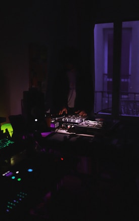 A dimly lit room with a person operating DJ equipment. Purple and blue lighting creates a moody atmosphere, and various electronic devices are visible, adding to the ambiance of a night-time music setup.