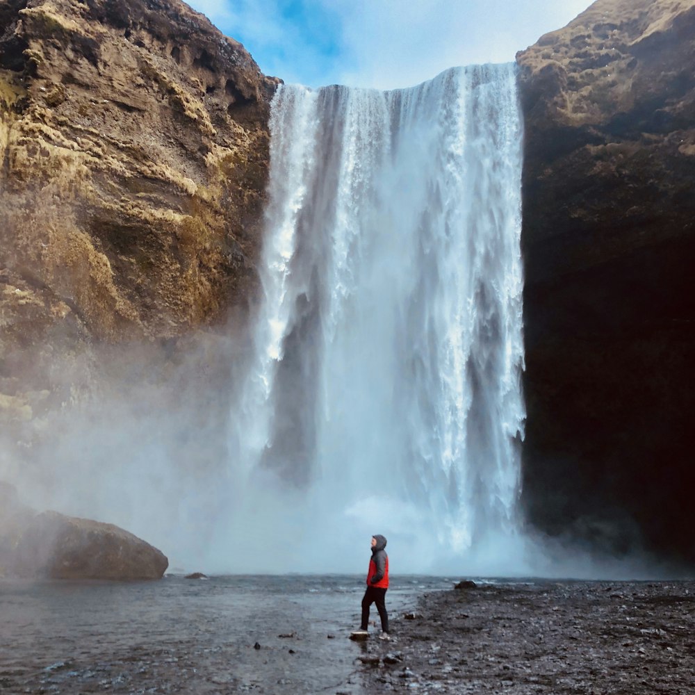 man in red jacket standing near waterfalls during daytime