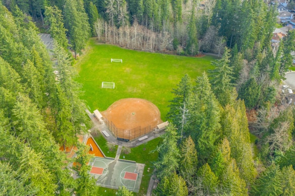 An aerial view of a sports complex surrounded by dense forest. The complex features a baseball diamond with an adjacent basketball court. The grass around the fields is lush and green, and two soccer goals are positioned on the field. The area around the complex is densely wooded, creating a serene and secluded environment.