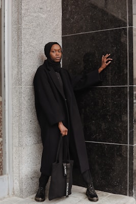 A person stands against a textured stone wall and a glossy black wall, wearing a black coat, headscarf, and boots. They hold a black tote bag with white text, and their pose exudes a sense of poise and confidence, with one hand resting on the black wall.