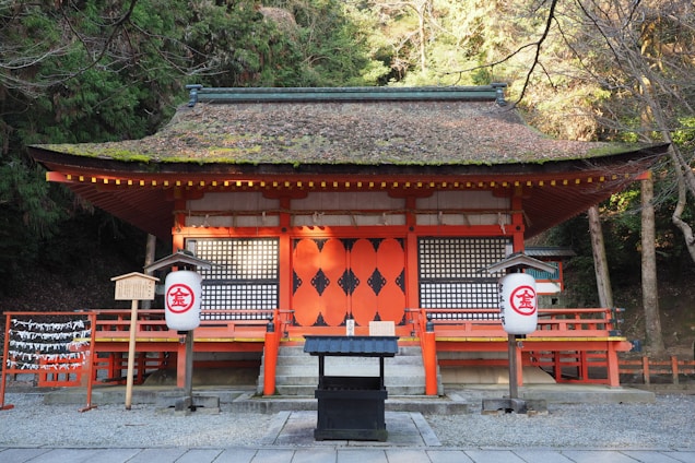 A traditional Japanese shrine featuring red-painted wooden structures with intricate lattice windows. The thatched roof is partially covered in moss, suggesting age and integration with nature. The shrine is adorned with large lanterns displaying red kanji characters. Surrounded by tall, dense trees, the scene evokes a sense of serenity and reverence. Stone steps lead up to the entrance, and wooden ema plaques are hung on the side, offering a place for visitors' wishes.