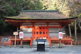 A traditional Japanese shrine featuring red-painted wooden structures with intricate lattice windows. The thatched roof is partially covered in moss, suggesting age and integration with nature. The shrine is adorned with large lanterns displaying red kanji characters. Surrounded by tall, dense trees, the scene evokes a sense of serenity and reverence. Stone steps lead up to the entrance, and wooden ema plaques are hung on the side, offering a place for visitors' wishes.