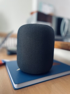 A black smart speaker with a sleek, mesh design sits on top of a closed blue notebook on a wooden table. The background is blurred, showing parts of a home interior, possibly a kitchen with light-colored walls and subtle hints of other objects.