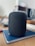 A black smart speaker with a sleek, mesh design sits on top of a closed blue notebook on a wooden table. The background is blurred, showing parts of a home interior, possibly a kitchen with light-colored walls and subtle hints of other objects.