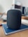 A black smart speaker with a sleek, mesh design sits on top of a closed blue notebook on a wooden table. The background is blurred, showing parts of a home interior, possibly a kitchen with light-colored walls and subtle hints of other objects.