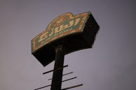 An elevated signboard with Arabic text is positioned against a dim, dusky sky. The sign features bright colors, including orange and green, with intricate designs at the top. The pole holding the sign is black and rusted, suggesting age or wear.