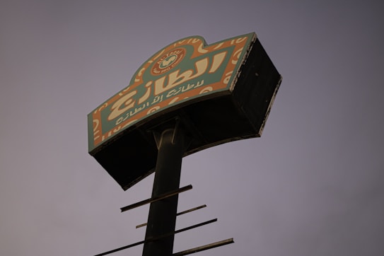 An elevated signboard with Arabic text is positioned against a dim, dusky sky. The sign features bright colors, including orange and green, with intricate designs at the top. The pole holding the sign is black and rusted, suggesting age or wear.