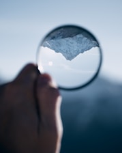 A hand holding a magnifying glass that inverts and magnifies the image of a snow-covered mountain peak against a clear blue sky.