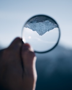 A hand holding a magnifying glass that inverts and magnifies the image of a snow-covered mountain peak against a clear blue sky.