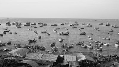 Numerous small boats are floating close to shore in a large body of water. The scene is vibrant with maritime activity, as people can be seen tending to the boats. Near the shore, market stalls with canopies are visible, suggesting a busy coastal marketplace.