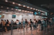 People are standing in line at a ticket counter, likely at a transportation hub such as a train station or airport. The counter displays various ticket options and information on overhead signs. The area is well-lit and organized, with barriers guiding the queues.