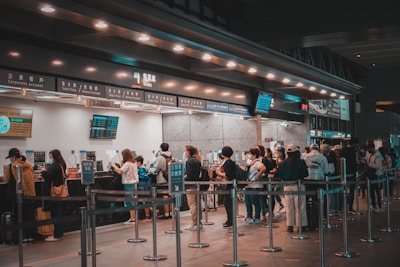People are standing in line at a ticket counter, likely at a transportation hub such as a train station or airport. The counter displays various ticket options and information on overhead signs. The area is well-lit and organized, with barriers guiding the queues.