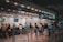 People are standing in line at a ticket counter, likely at a transportation hub such as a train station or airport. The counter displays various ticket options and information on overhead signs. The area is well-lit and organized, with barriers guiding the queues.
