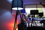 A moody indoor scene featuring a modern workspace. There are two tall black lamps with a soft glow, one casting blue light and the other casting a warm orange hue. In the background, a desk with multiple screens, a chair, and various electronics can be seen. Daylight filters in through a large window, with faint greenery visible outside. Decor and objects, such as a bicycle wheel and a set of headphones, add to the eclectic vibe.
