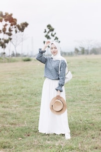 woman in white long sleeve dress holding brown hat standing on green grass field during daytime