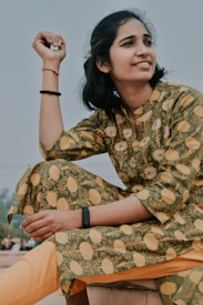 A person sits outdoors, wearing a green and yellow patterned traditional outfit. The individual is smiling and looking to the side, with a relaxed and content expression. They have a black fitness band on their wrist.